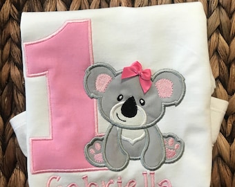 Könnte beinhalten: Ein weißer Strampler mit einer pinkfarbenen Zahl eins und einem grauen Koala mit einer pinkfarbenen Schleife. Der Strampler ist mit dem Namen "Gabriella" personalisiert.