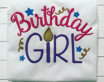 Puede incluir: Una camiseta blanca con texto bordado que dice Birthday en rosa y GIRL en morado. Una llama de vela con purpurina dorada se sitúa sobre la letra I de GIRL, mientras que estrellas azules y espirales doradas rodean las letras.