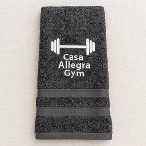 May include: A dark gray hand towel featuring an embroidered white barbell icon above the text Casa Allegra Gym. The towel has a textured terry cloth surface with two horizontal decorative bands near the bottom edge.