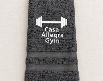 May include: A dark gray hand towel featuring an embroidered white barbell icon above the text Casa Allegra Gym. The towel has a textured terry cloth surface with two horizontal decorative bands near the bottom edge.