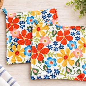 May include: Two square fabric pot holders featuring a white background with a vibrant floral pattern in shades of red, yellow, pink, and blue, accented by green leaves and stems. Each pot holder includes a small blue fabric hanging loop.