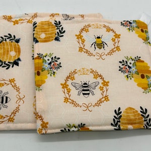 May include: Two beige pot holders with a bee and honeycomb pattern. The pot holders have a white loop for hanging.