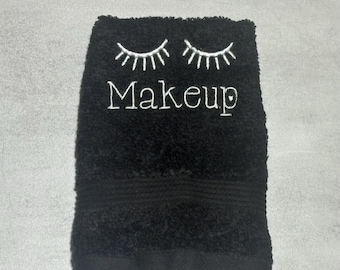 May include: A black rectangular hand towel featuring white embroidered text that reads Makeup, positioned below a pair of stylized white embroidered eyelashes. The towel has a textured terry cloth surface and a smooth hemmed border at the bottom.