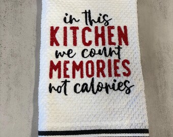 May include: A white waffle-weave kitchen towel featuring the black and red embroidered text: in This KITCHEN we count MEMORIES not calories. The bottom of the towel is decorated with three horizontal black stripes against a white background.