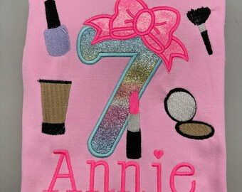 Puede incluir: Una camiseta rosa claro con un diseño bordado de un gran número siete arcoíris brillante rematado con un lazo rosa intenso. Alrededor del número hay artículos de maquillaje bordados que incluyen esmalte de uñas, brocha, pintalabios y polvos.