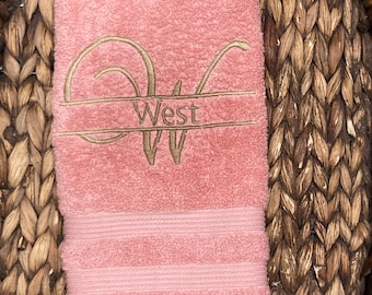 May include: A pink hand towel with a gold embroidered monogram of the letter "W" and the name "West" above it.