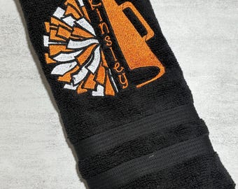 Puede incluir: Una toalla de mano negra con un diseño bordado de un megáfono naranja y un pompón en naranja y blanco. El nombre KINSLEY está bordado verticalmente en letras naranjas a lo largo del lado del megáfono sobre la tela texturizada.
