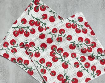 Puede incluir: Dos servilletas de tela cuadradas con fondo blanco y un patrón de cuadros gris claro. La tela está decorada con un estampado repetitivo de tomates rojos sobre tallos y hojas verdes, dispuestos de forma uniforme por toda la superficie.