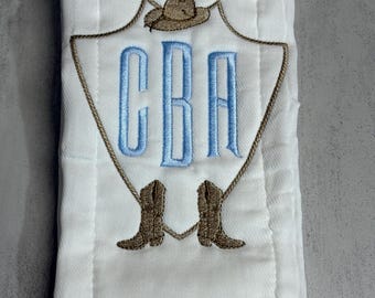 May include: A white burp cloth featuring an embroidered design of a brown cowboy hat at the top and two brown cowboy boots at the bottom, connected by a rope outline. In the center, the letters CBA are embroidered in light blue thread.