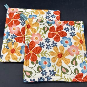 May include: Two white cotton pot holders with a floral pattern in shades of red, yellow, blue, and green. The pot holders have a light blue trim around the edges.