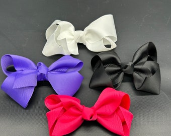 May include: Four hair bows made of ribbed fabric, each featuring a central knot and looped ends. The set includes one white bow, one purple bow, one black bow, and one red bow, all arranged in a cluster against a dark, textured background.