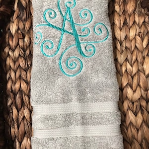 May include: A gray hand towel with a teal embroidered letter "A" in a decorative script. The towel is folded and placed in a woven basket. The towel has a textured surface and white stripes near the bottom.