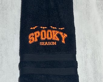 May include: A black hand towel featuring an embroidered design in orange thread. The embroidery includes four small flying bats positioned above the text SPOOKY SEASON in a bold, collegiate-style font. The towel has a textured terry cloth finish.