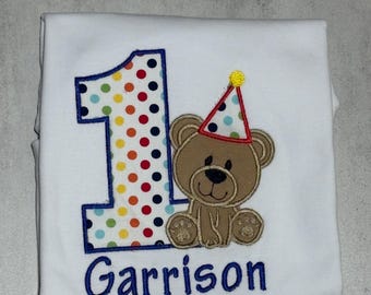 Puede incluir: Una camiseta blanca con un diseño bordado de un número uno grande relleno de lunares multicolores, un oso de peluche marrón con un sombrero de fiesta de lunares de colores y el nombre Garrison bordado en hilo azul debajo del gráfico.