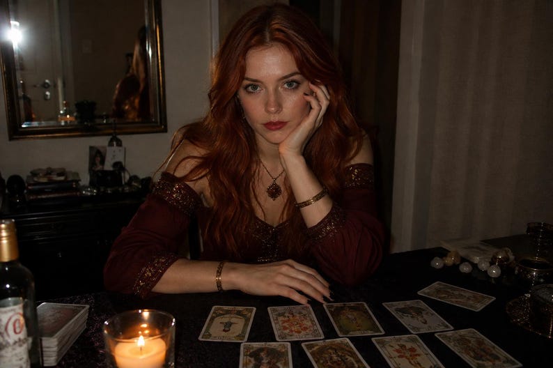 May include: A person with long red hair wearing a dark maroon off-the-shoulder top with gold-toned metallic trim on the sleeves and neckline. The person wears a gold-toned necklace and bracelets while sitting at a table with several tarot cards.