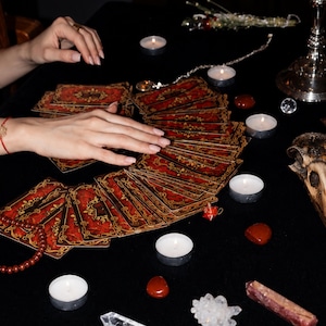 May include: A deck of tarot cards with red and gold ornate patterns spread out in a fan shape. The cards are surrounded by several small white tea light candles, polished red stones, a crystal cluster, a clear crystal point, and a skull.