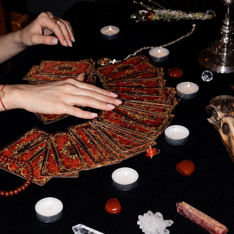 May include: A deck of tarot cards with red and gold ornate patterns spread out in a fan shape. The cards are surrounded by several small white tea light candles, polished red stones, a crystal cluster, a clear crystal point, and a skull.