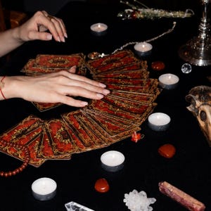 K&ouml;nnte beinhalten: Ein Tarotkartendeck mit verzierten roten und goldenen Mustern, das f&auml;cherf&ouml;rmig ausgebreitet ist. Die Karten sind umgeben von mehreren kleinen wei&szlig;en Teelichtern, polierten roten Steinen, einer Kristallgruppe, einer Kristallspitze und einem Sch&auml;del.