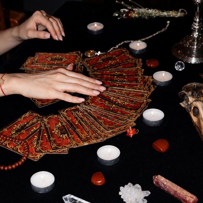 K&ouml;nnte beinhalten: Ein Tarotkartendeck mit verzierten roten und goldenen Mustern, das f&auml;cherf&ouml;rmig ausgebreitet ist. Die Karten sind umgeben von mehreren kleinen wei&szlig;en Teelichtern, polierten roten Steinen, einer Kristallgruppe, einer Kristallspitze und einem Sch&auml;del.