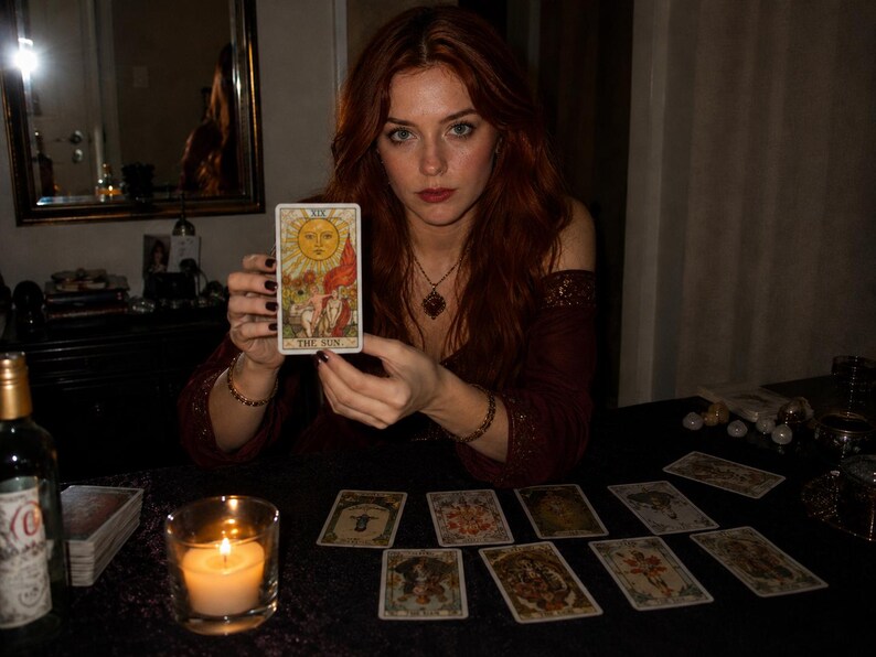 K&ouml;nnte beinhalten: Eine rechteckige Tarotkarte mit dem Titel The Sun, die eine gelbe Sonnenillustration &uuml;ber einem Kind auf einem wei&szlig;en Pferd zeigt, gehalten von einer Person in einem dunkelroten schulterfreien Oberteil mit goldener Halskette und Armb&auml;ndern.