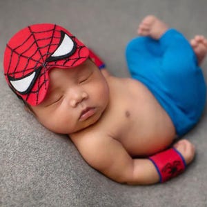 Puede incluir: Un bebé recién nacido vestido con un disfraz de Spider-Man, que incluye una máscara roja y negra con detalles blancos en los ojos, pantalones azules y muñequeras rojas. El bebé está acostado sobre una superficie gris.