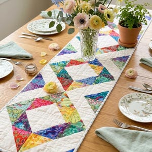 May include: A colorful quilted table runner with a geometric pattern of triangles in vibrant hues of red, blue, yellow, and green. The runner is placed on a wooden table, alongside a vase of flowers, plates, and silverware.