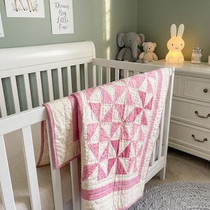 以下が含まれることがあります： ピンクと白のキルトが側面に掛けられた白いベビーベッド。キルトは幾何学的なスターバースト模様です。保育園には、灰色のラグ、ぬいぐるみ、鹿と月のウォールアートも含まれています。