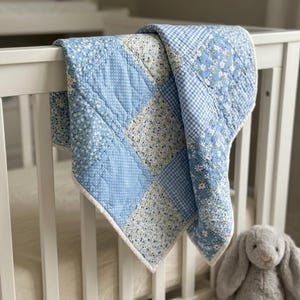 Puede incluir: Un edredón de bebé patchwork en tonos azules, blancos y crema, sobre una cuna blanca. El edredón presenta varios patrones, incluyendo flores, lunares y cuadros. Un conejo de peluche gris está junto a la cuna.