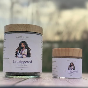 May include: Two glass jars of herbal tea with wooden lids. The labels read "Anita Herbs" and "Untriggered Herbal Tea." The larger jar is labeled 8oz, and the smaller jar is labeled 2oz. Each label features an illustration of a woman.