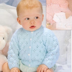 Può includere: Un cardigan di maglia blu chiaro per neonato con chiusura a bottoni e un delicato motivo a pizzo. Il cardigan è indossato da un bambino seduto su una coperta bianca.