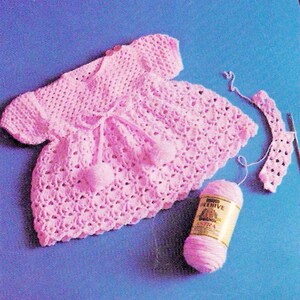 May include: A light pink crocheted dress with short sleeves and a tie at the waist, featuring two small pom-poms. A ball of pink yarn and a crocheted piece are also visible. The text "Girls Boys Dolls N Toys" is at the top.