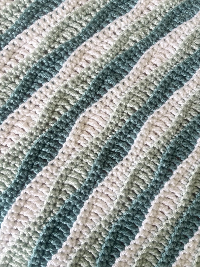 May include: A close-up of a crocheted blanket with diagonal stripes. The stripes alternate between white, light green, and a darker teal green. The texture of the crochet stitches is visible, creating a soft, textured appearance.