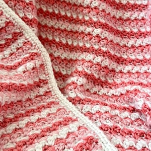 May include: A close-up of a pink and white crocheted blanket with a textured pattern. The blanket is folded in half, showing the different colors and textures.