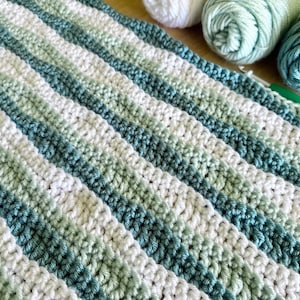 May include: Crocheted blanket with diagonal stripes in shades of white, green, and teal. The handmade blanket is made with soft yarn. The image also shows several balls of yarn in coordinating colors, and a crochet hook.
