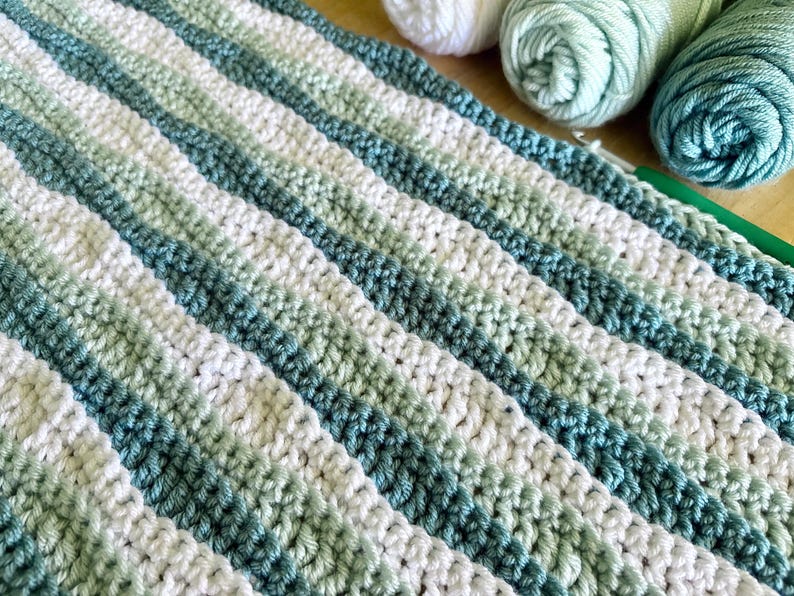 May include: Crocheted blanket with diagonal stripes in shades of white, green, and teal. The handmade blanket is made with soft yarn. The image also shows several balls of yarn in coordinating colors, and a crochet hook.