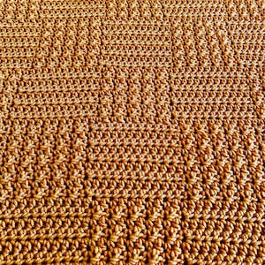 Puede incluir: Primer plano de una manta de ganchillo hecha a mano en un cálido tono marrón dorado. La superficie texturizada muestra intrincados patrones de puntadas, creando un textil denso y visualmente atractivo. El color y el diseño de la manta sugieren un artículo acogedor y artesanal.