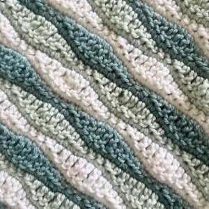 May include: Close-up of a handmade crochet blanket with diagonal stripes. The blanket features alternating stripes of white, light green, and teal blue yarn. The texture of the crochet stitches is visible, creating a cozy and textured appearance.