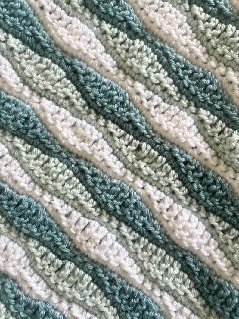 May include: Close-up of a handmade crochet blanket with diagonal stripes. The blanket features alternating stripes of white, light green, and teal blue yarn. The texture of the crochet stitches is visible, creating a cozy and textured appearance.