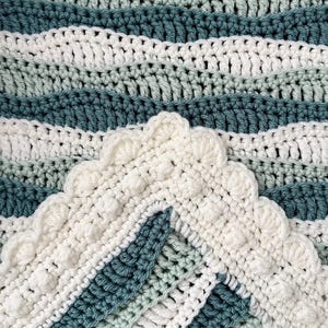 May include: A handmade crochet blanket in shades of white, teal, and light green. The blanket features a wavy pattern and a decorative scalloped edge. The yarn appears soft and the stitches are tightly woven.