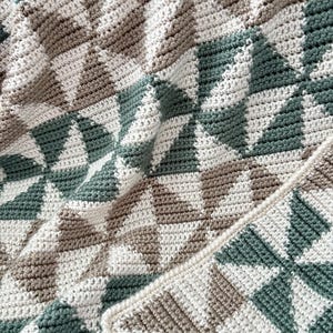May include: A hand-crocheted blanket with a geometric design in cream, sage green, and brown. The pattern consists of repeating diamond shapes, giving the blanket a textured appearance. The colour scheme is neutral, making it suitable for different interior design styles. The blanket is approximately 150cm x 200cm.