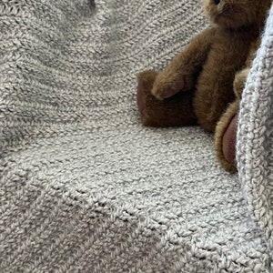 May include: A grey knitted throw blanket with a brown teddy bear sitting on top of it.