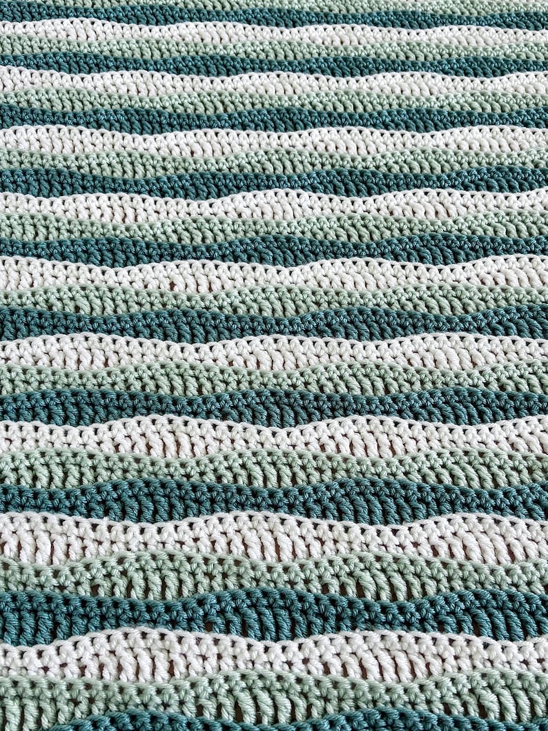 May include: A handmade crochet blanket with a wave-like pattern. The blanket features alternating stripes of white, light green, medium green, and teal. The texture is visible, showcasing the individual stitches.