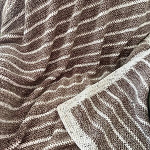 May include: A crocheted blanket featuring alternating horizontal stripes of dark brown and cream yarn. The texture is created with a consistent stitch pattern throughout the fabric, including a wider cream-colored border along the edge.