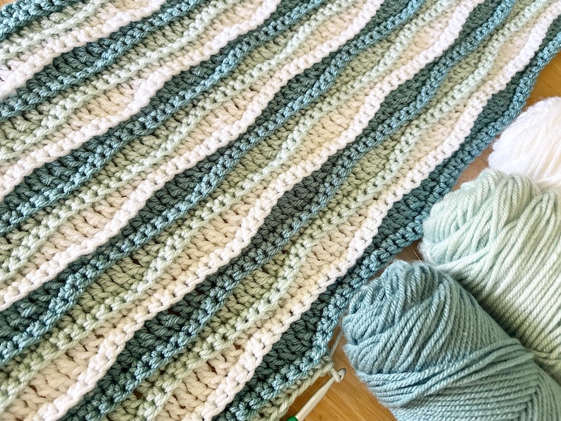 May include: A handmade crochet blanket in shades of blue, green, and white, with a wavy striped pattern. Several balls of yarn in matching colors are visible, suggesting a crafting project. The blanket is made with a crochet hook.