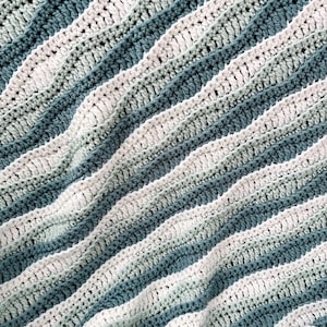 May include: A close-up of a handmade crochet blanket. The blanket features alternating stripes of white, light green, and teal blue. The textured stitches create a wavy pattern, adding visual interest to the soft, cozy textile.