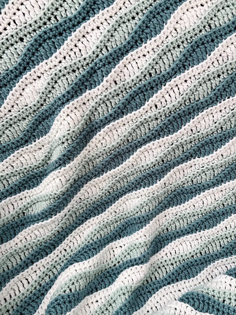 May include: A close-up of a handmade crochet blanket. The blanket features alternating stripes of white, light green, and teal blue. The textured stitches create a wavy pattern, adding visual interest to the soft, cozy textile.