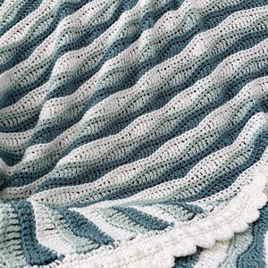May include: A handmade crochet blanket in shades of white, teal, and blue. The blanket features a wavy, striped pattern and a scalloped edge. The soft, textured design is perfect for home decor.