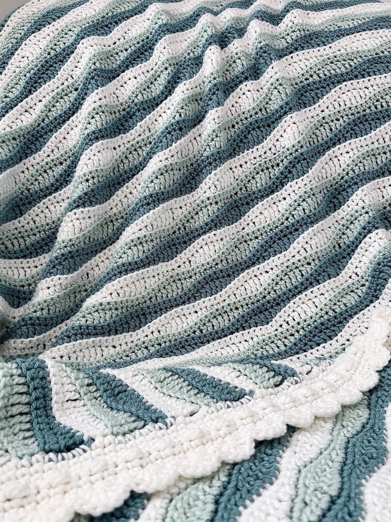 May include: A handmade crochet blanket in shades of white, teal, and blue. The blanket features a wavy, striped pattern and a scalloped edge. The soft, textured design is perfect for home decor.