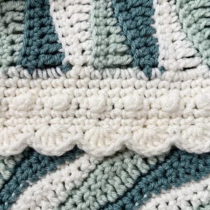 May include: Close-up of a handmade crochet blanket with a textured design. The blanket features alternating stripes of white, teal, and light green. A scalloped edge and bobble stitch detail add visual interest to this cozy, handcrafted item.
