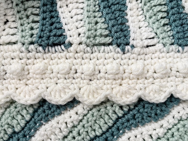 May include: Close-up of a handmade crochet blanket with a textured design. The blanket features alternating stripes of white, teal, and light green. A scalloped edge and bobble stitch detail add visual interest to this cozy, handcrafted item.